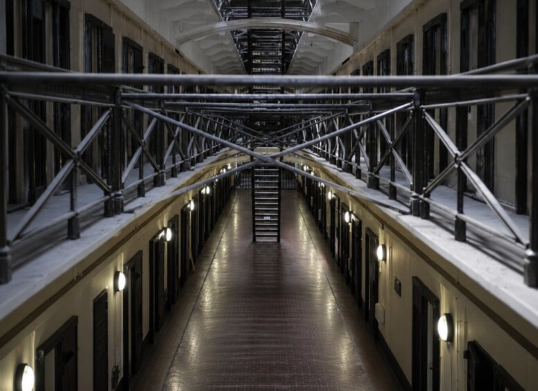 A long, narrow corridor with metal railings and doors on both sides, leading to a staircase in the center. 4