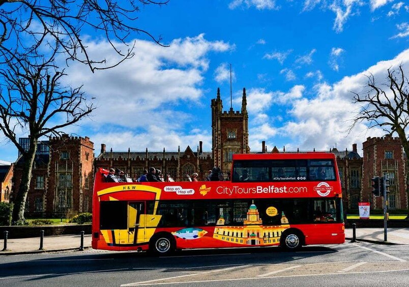 A red double-decker bus with 'CityToursBelfast.com' and 'Hop on Hop off' logos, parked in front of a historic building with a clock tower. 5