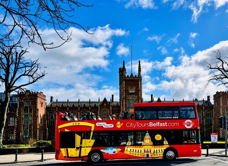 A red double-decker bus with 'CityToursBelfast.com' and 'Hop on Hop off' on its side, parked in front of a historic building with a clock tower. 1