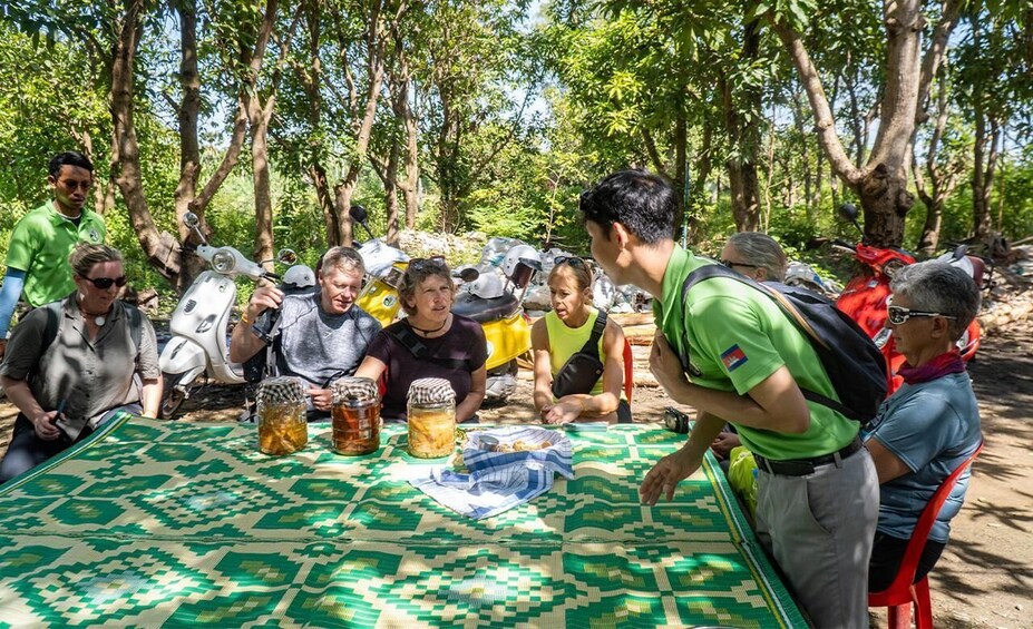 Un groupe de personnes est assis autour d’une table recouverte d’une nappe à motifs verts, et profite d’un repas en plein air dans un endroit boisé. 5