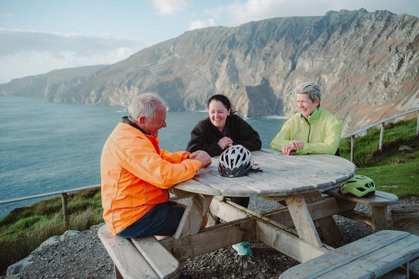 Tres personas sentadas en una mesa de picnic con un casco encima, con vistas a un paisaje costero y montañas al fondo. 3