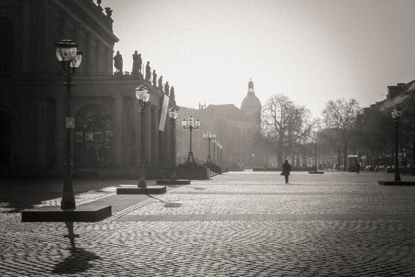 A cobblestone street with a person walking, buildings with statues, and a domed structure in the distance. 3