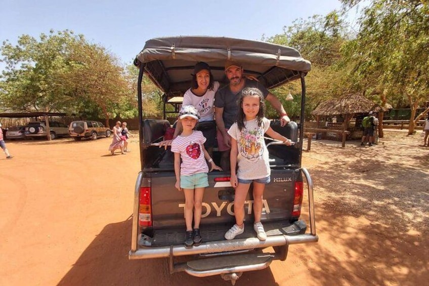 A Toyota SUV with a roof, a family of four, and a dirt ground with trees and a wooden structure in the background. 4