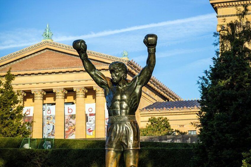 En bronze statue af en boksertype med løftede arme foran en klassisk bygning med søjler. 1
