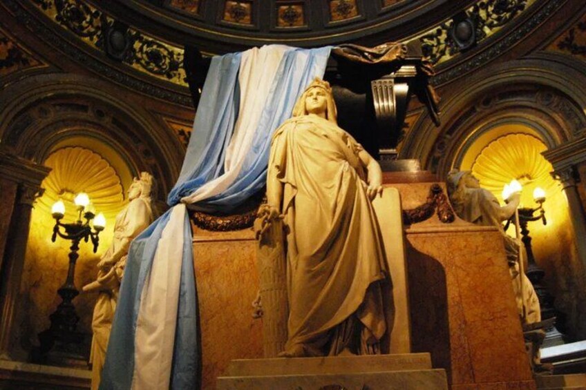A statue of a woman in classical attire, draped in blue and white cloth, standing on a pedestal in a grand hall with ornate architecture and two lit lamps on either side. 2