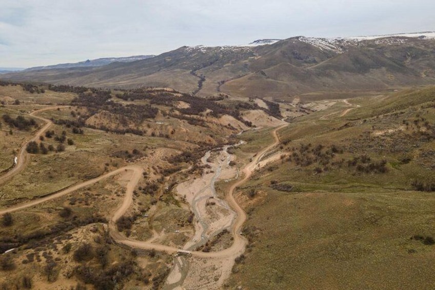 Un chemin de terre sinueux traverse un paysage vallonné, où la végétation est clairsemée et où l’on aperçoit, au loin, des montagnes aux sommets enneigés. 2