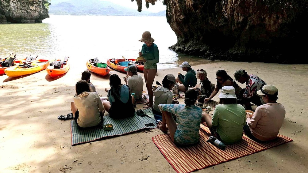 A group of people sitting on mats on a sandy beach, with kayaks lined up in the background. 5