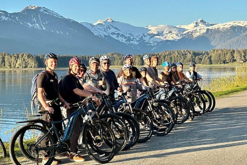 A group of cyclists on a road with a lake and mountains in the background. 1