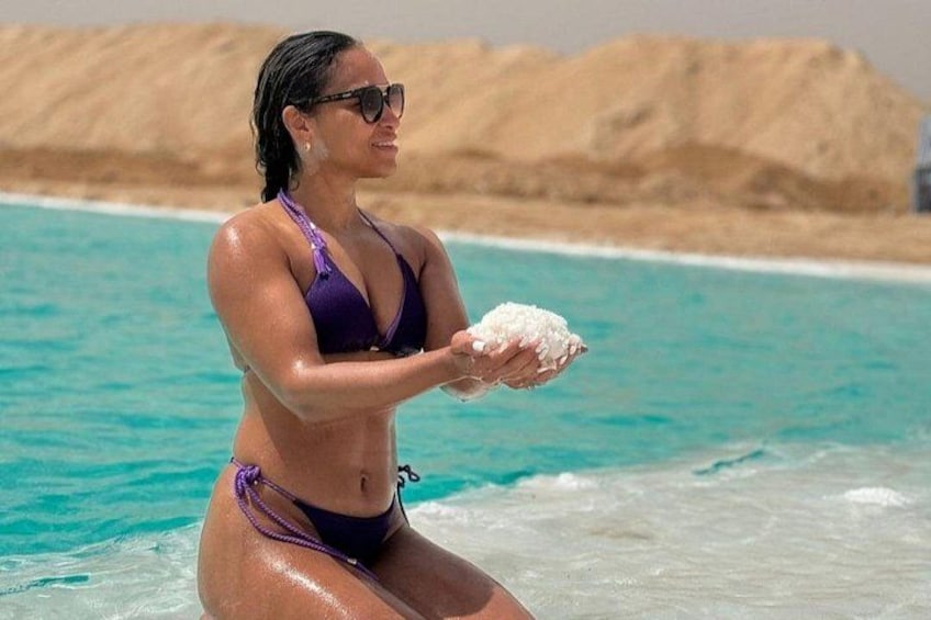 A person in a purple bikini holding a shell at a beach with clear turquoise water and a sandy shore. 1