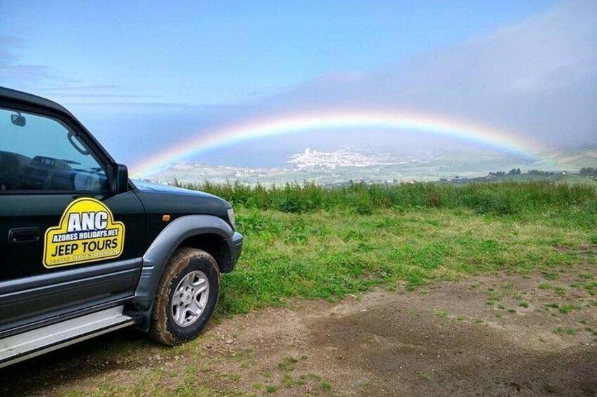 Un véhicule arborant le logo « ANC Azores Holidays & Jeep Tours » est garé sur un chemin de terre, avec un champ herbeux et un arc-en-ciel en arrière-plan. 5