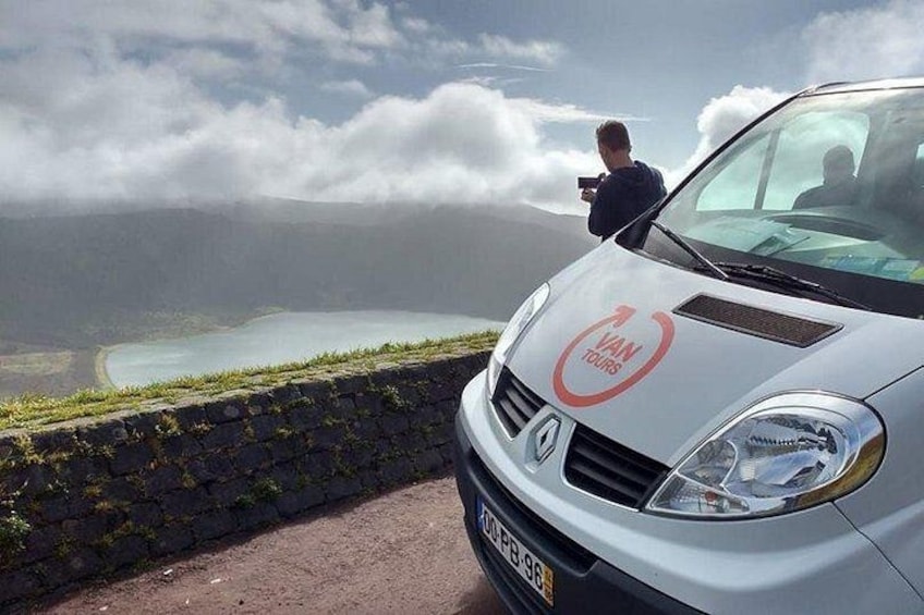 A white van with 'VAN TOURS' on the front, parked on a road with a stone wall and a view of a lake and mountains in the background. 5