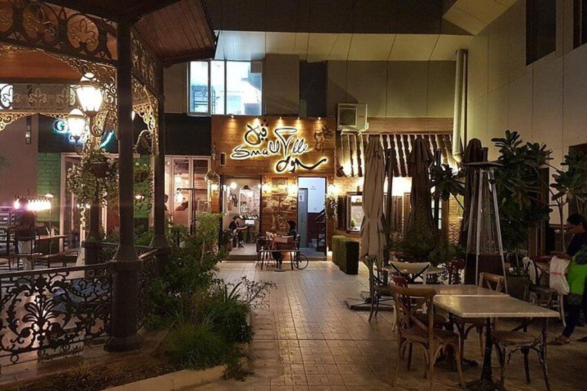 An outdoor cafe area with tables and chairs, a neon sign, and potted plants. 2