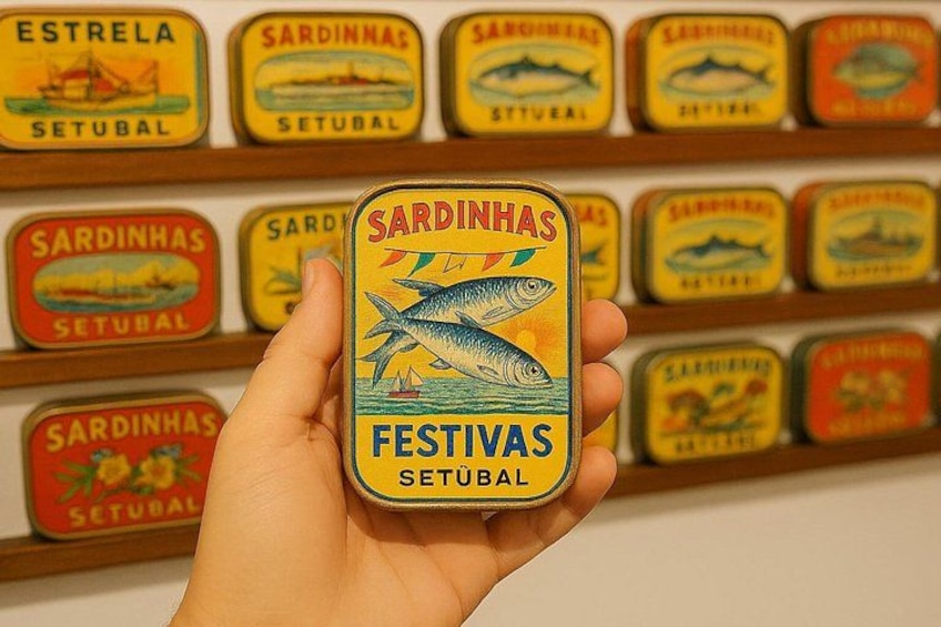 Eine Hand hält eine Dose mit dem Aufdruck 'Sardinhas Festivas Setubal', im Hintergrund eine Reihe ähnlicher Dosen. 1