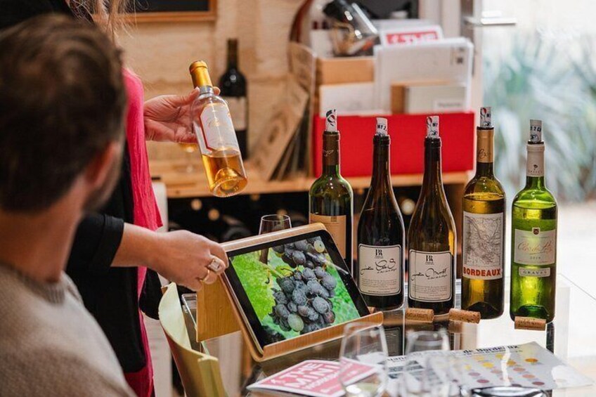 A person holding a wine bottle, a tablet displaying grapes, and several wine bottles on a table. 4