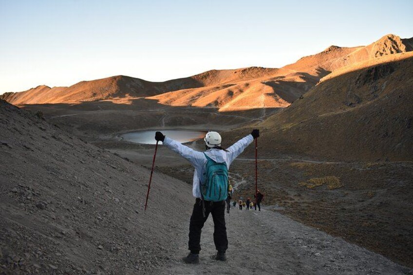 A person with arms raised, standing on a dirt path, facing a mountainous landscape with a lake in the distance. 4