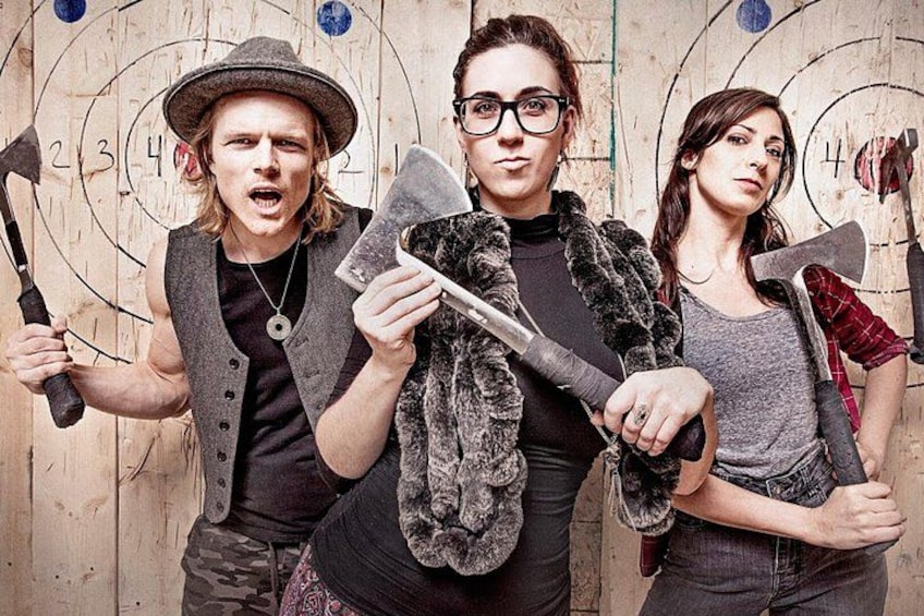 Three people holding axes in front of a wooden board with numbered circles. 1