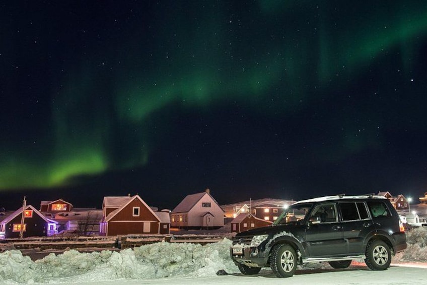 A black SUV parked in a snowy area with houses in the background and the Northern Lights in the sky. 2
