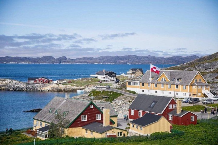 A coastal village with houses, a flag, and a view of the sea and mountains. 1
