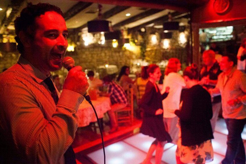 Een man zingt in de microfoon in een donkere locatie, met een publiek op de achtergrond. 4
