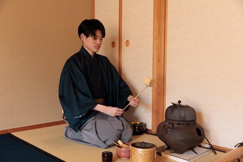 Een persoon in traditionele kledij zittend op een tatami-mat, een hamer vasthoudend, met een theeketel en andere voorwerpen op een tafel. 3