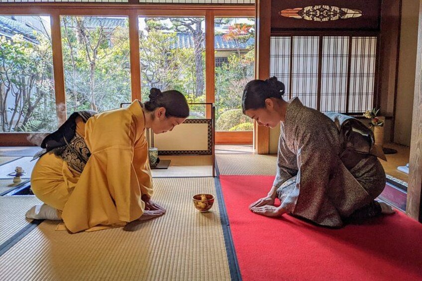 Deux personnes, vêtues de costumes traditionnels, effectuent un rituel sur des tatamis dans une pièce d’où l’on peut admirer des arbres et dont l’intérieur est de style japonais traditionnel. 2