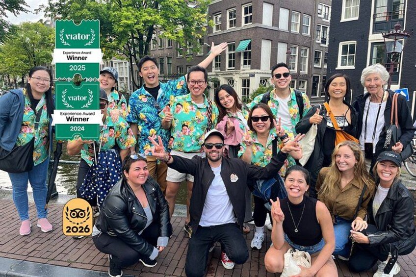 Een groep mensen poseert met een bordje 'Visitor Award Winner', op een stedelijke locatie met gebouwen en een straat van bakstenen. 1