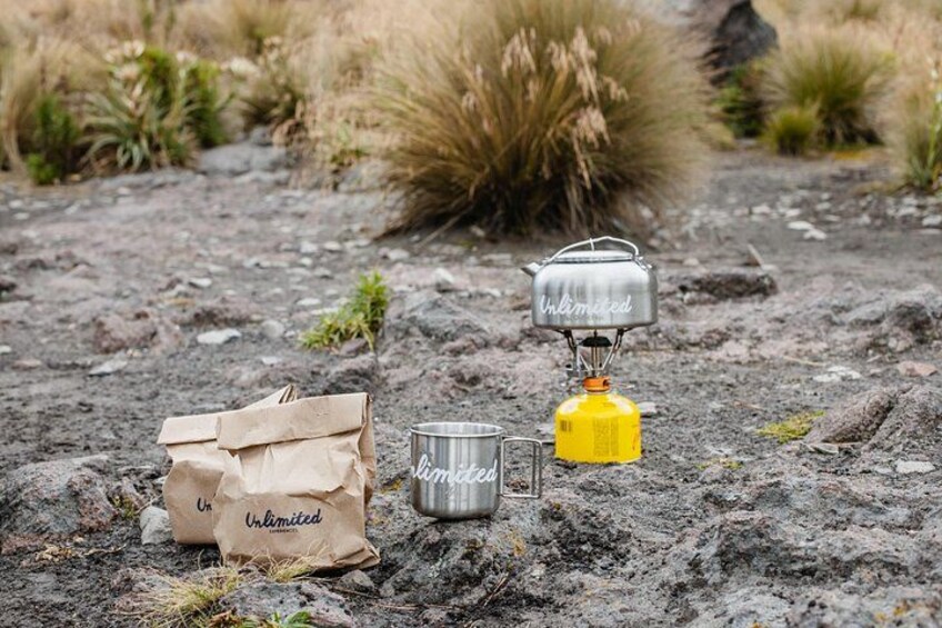 A camping setup with a metal pot, a yellow gas canister, and a paper bag labeled 'Unlimited' on a rocky ground. 4