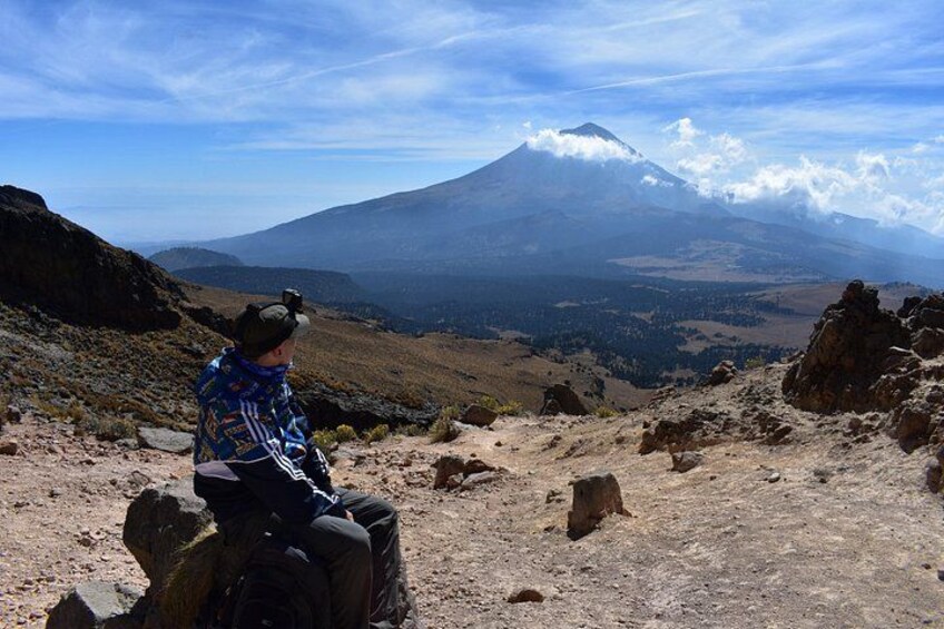 A person sitting on a rocky outcrop overlooking a mountainous landscape with a prominent peak. 3