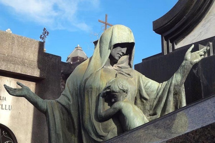 Estatua de una figura vestida con los brazos extendidos, una cruz al fondo y un edificio con inscripciones. 3