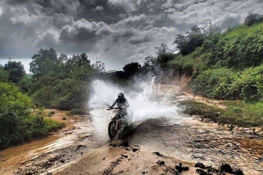 A person on a motorcycle, water splashing around them, on a muddy path with greenery on either side. 1