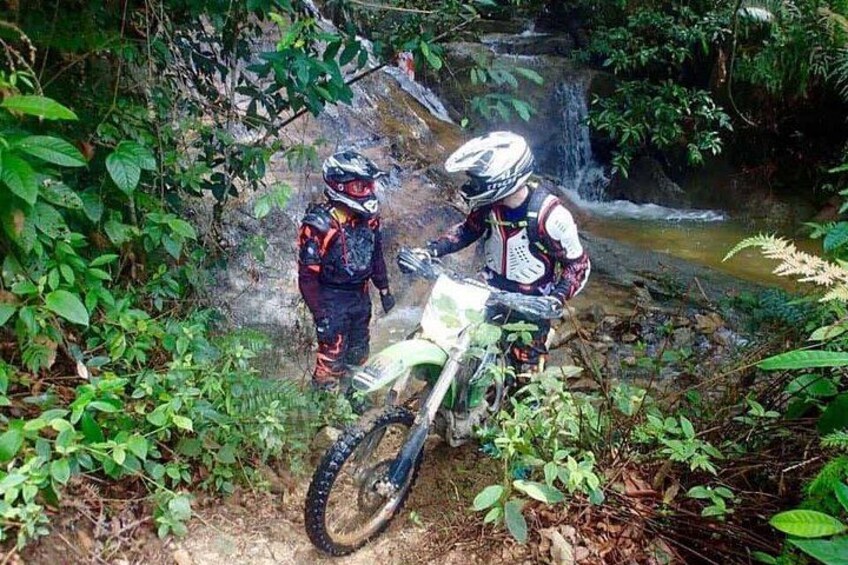 Two motorcyclists in protective gear on a dirt trail with a waterfall in the background. 3