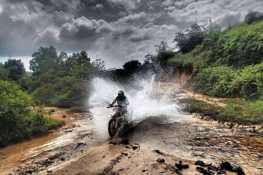 A person on a motorcycle creating a water spray on a dirt path surrounded by greenery. 5
