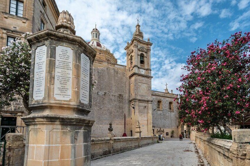 Un edificio histórico con un campanario, un monumento de piedra con inscripciones y un árbol en flor. 2