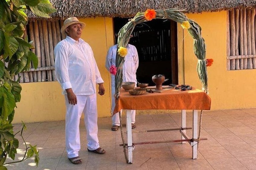 Een man staat voor een traditionele woning met rieten dak, een tafel met rituele objecten en een persoon op de achtergrond. 4