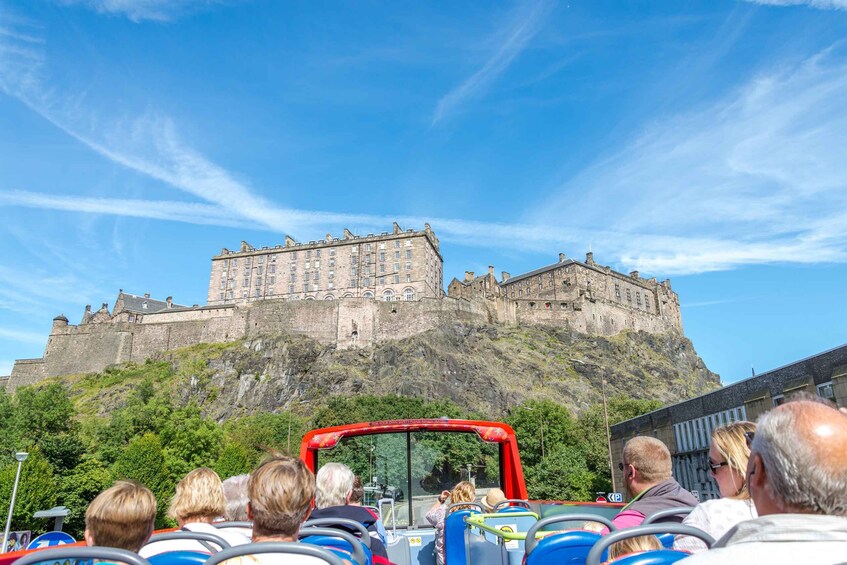 A group of tourists on a red bus, facing a historic castle on a hill. 2