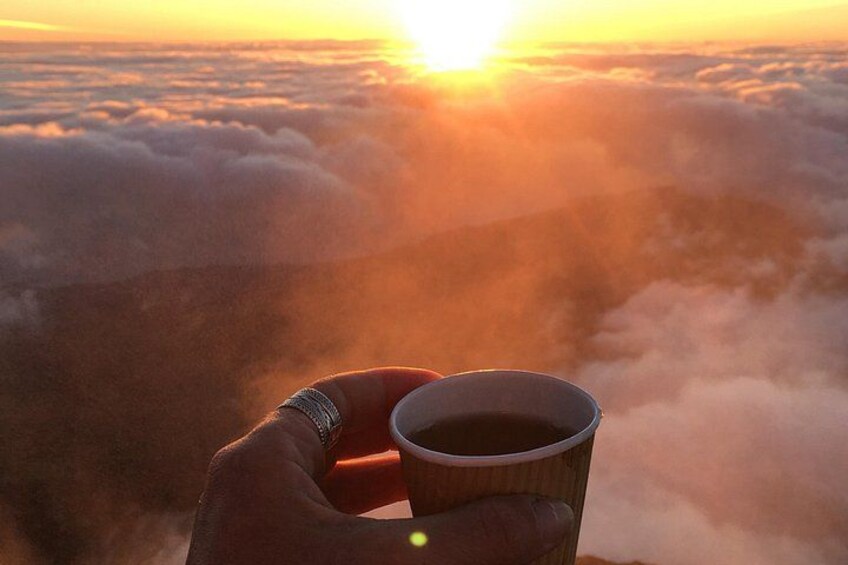 A hand holding a cup with a beverage, with a sunrise over clouds and mountains in the background. 5