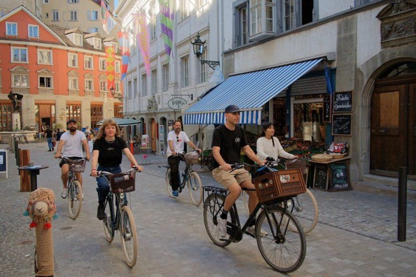 Folk cykler på en brostensbelagt gade med bygninger og flag. 1