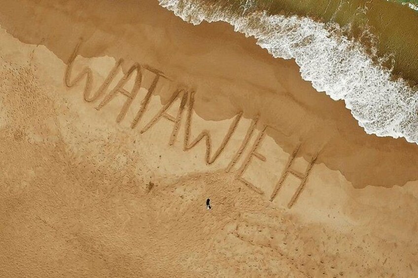 A sandy beach with the word 'WATANWEH' written in the sand and a small figure of a person. 2