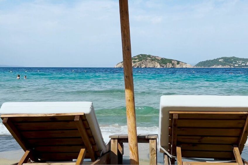 Two wooden lounge chairs facing a calm sea with clear blue water and a rocky island in the background. 4