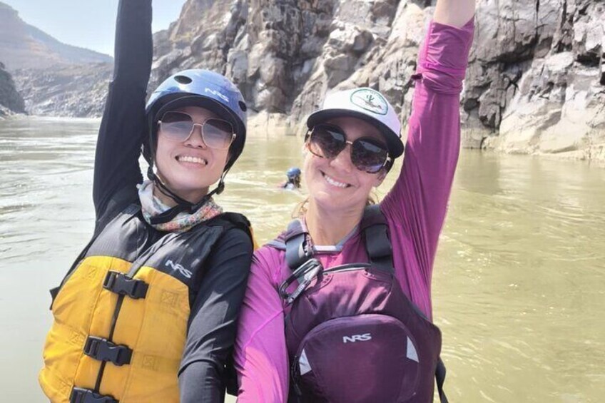 Two people wearing helmets and life jackets, standing in a river with rocky cliffs in the background. 2
