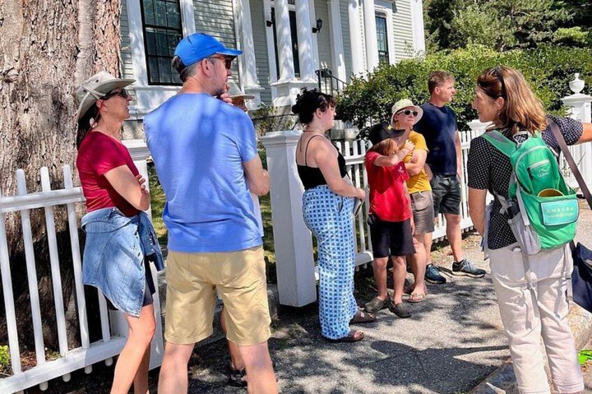 Eine Gruppe von Menschen steht draußen in der Nähe eines weißen Zauns, einige tragen Hüte und Sonnenbrillen, und eine Person hat einen grünen Rucksack dabei. 2