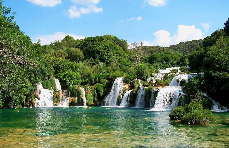 A series of waterfalls cascading into a clear, turquoise lake surrounded by lush greenery and hills. 1