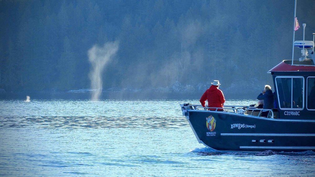 A boat with two people on it, a person in a red jacket and another in a blue jacket, with a whale spouting water in the background. 2