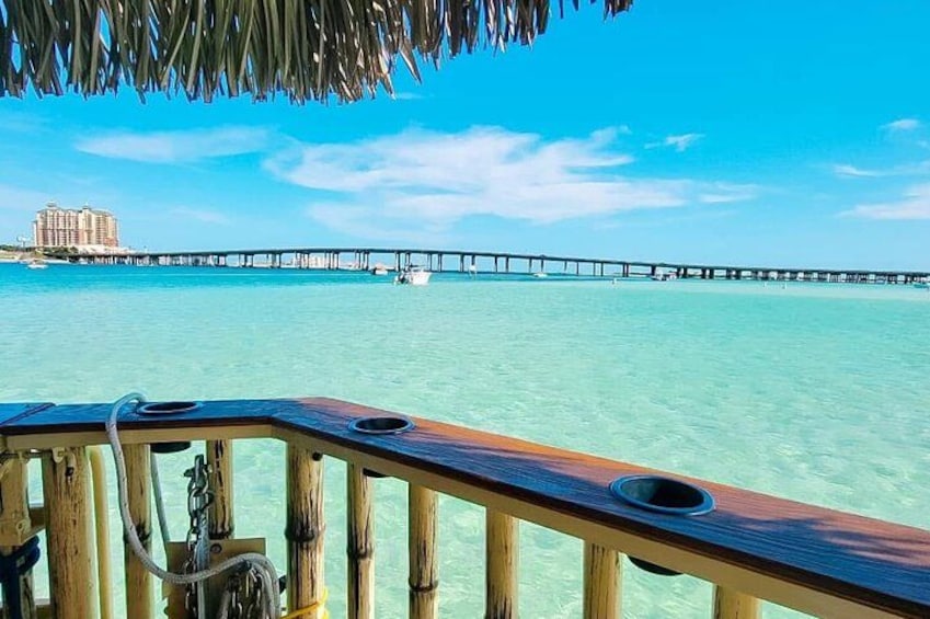 A wooden deck with a railing, a clear blue sky, turquoise water, and a pier in the distance. 1