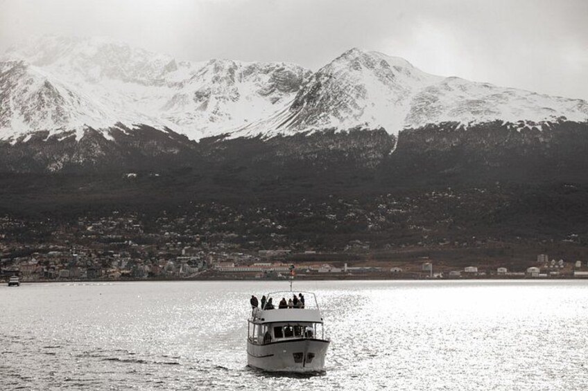 Ein Boot mit Passagieren auf ruhigem Gewässer, im Hintergrund ein schneegeb deckter Berg und eine Stadt am gegenüberliegenden Ufer. 5
