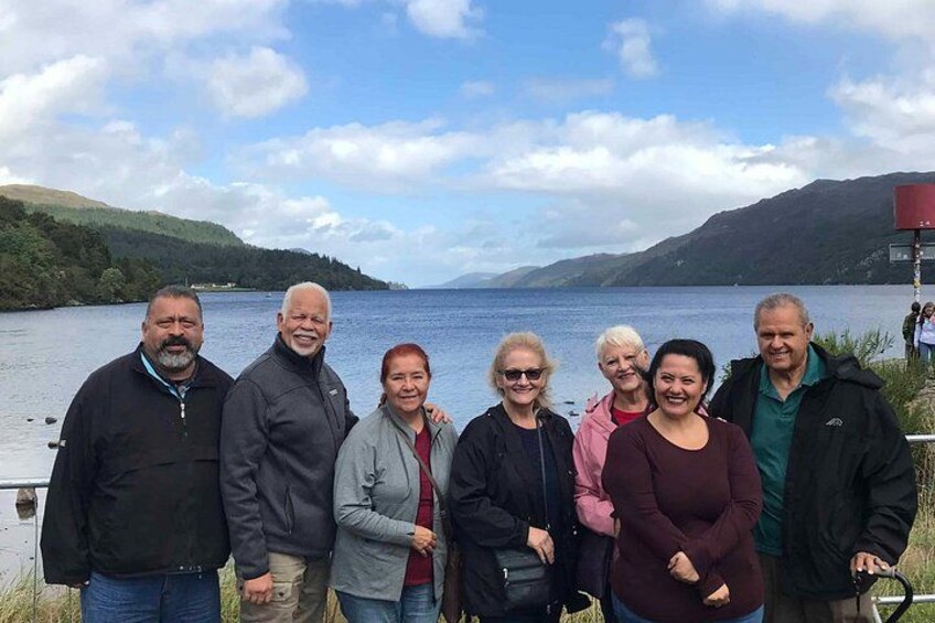 Un groupe de personnes se tient près d’un lac, avec des collines en arrière-plan. 1
