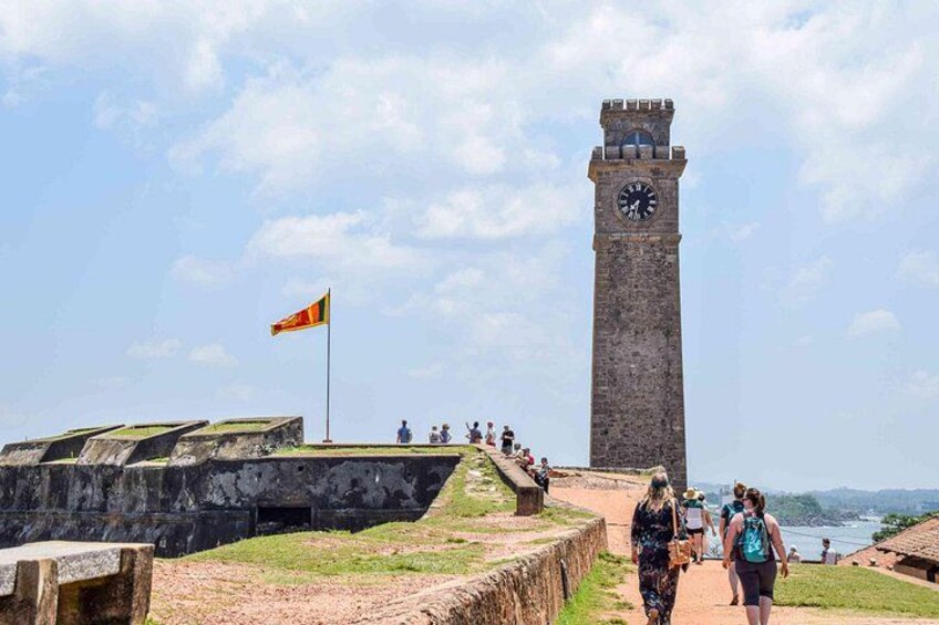 Een historische toren met een klok, een vlag en een vestingmuur. 1