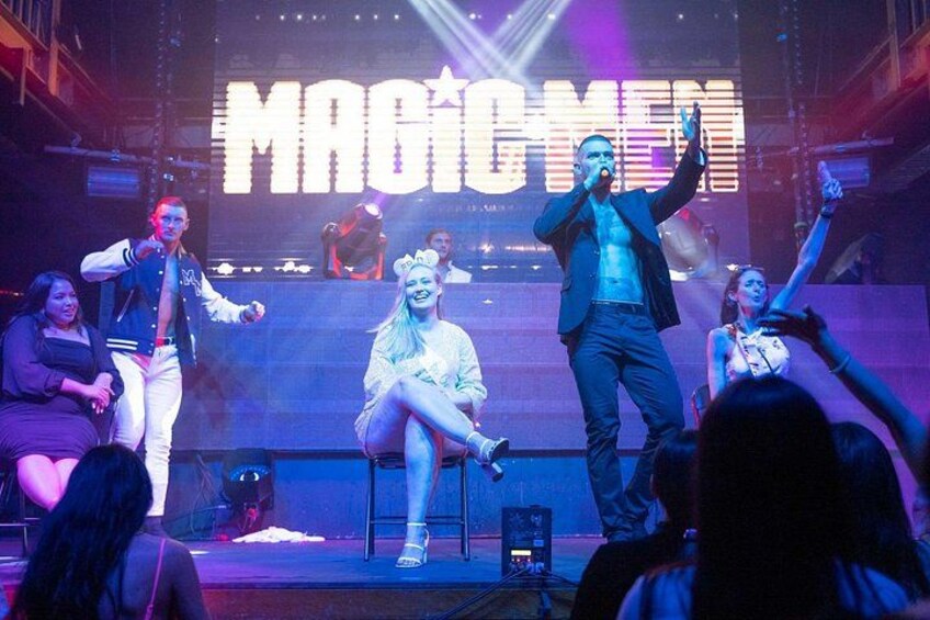 Un palco con artisti, un grande cartello illuminato con la scritta 'MAGIC MEN' e un pubblico. 5