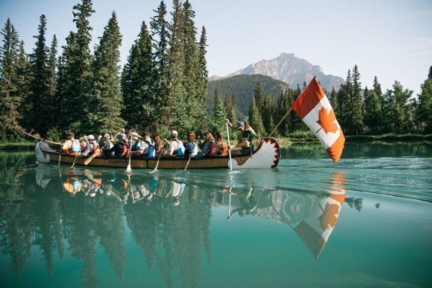 Een groep mensen in een kano op een kalm meer, met een Canadese vlag, omgeven door dicht bos en bergen op de achtergrond. 1