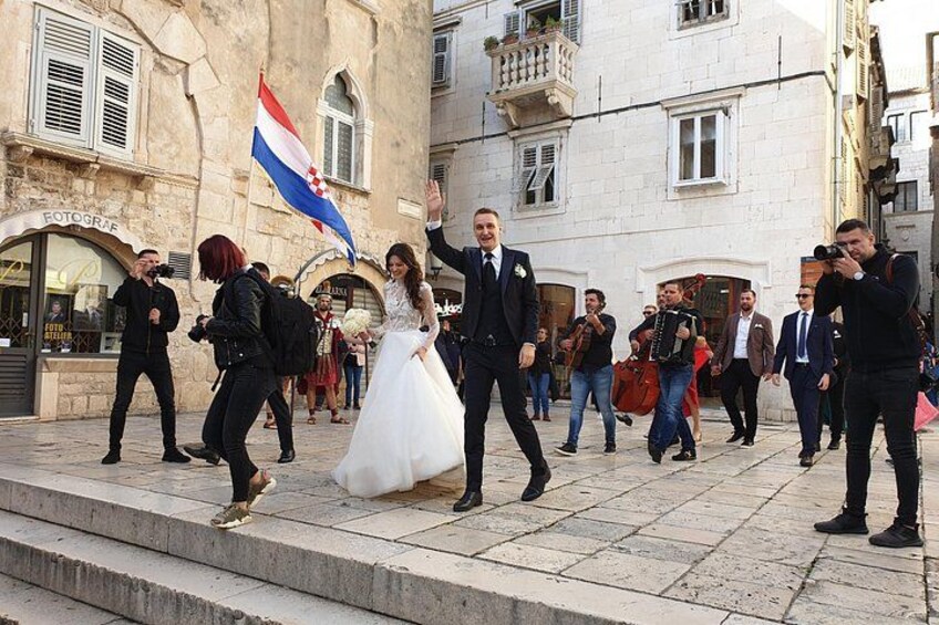 A wedding procession with a bride in a white dress and a groom in a black suit, walking down a stone-paved street. 2